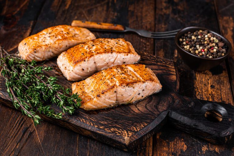 Three grilled salmon fillets seasoned with herbs and spices, served on a rustic wooden board alongside a bowl of mixed peppercorns and some fresh thyme sprigs, creating an appetizing and flavorful presentation.