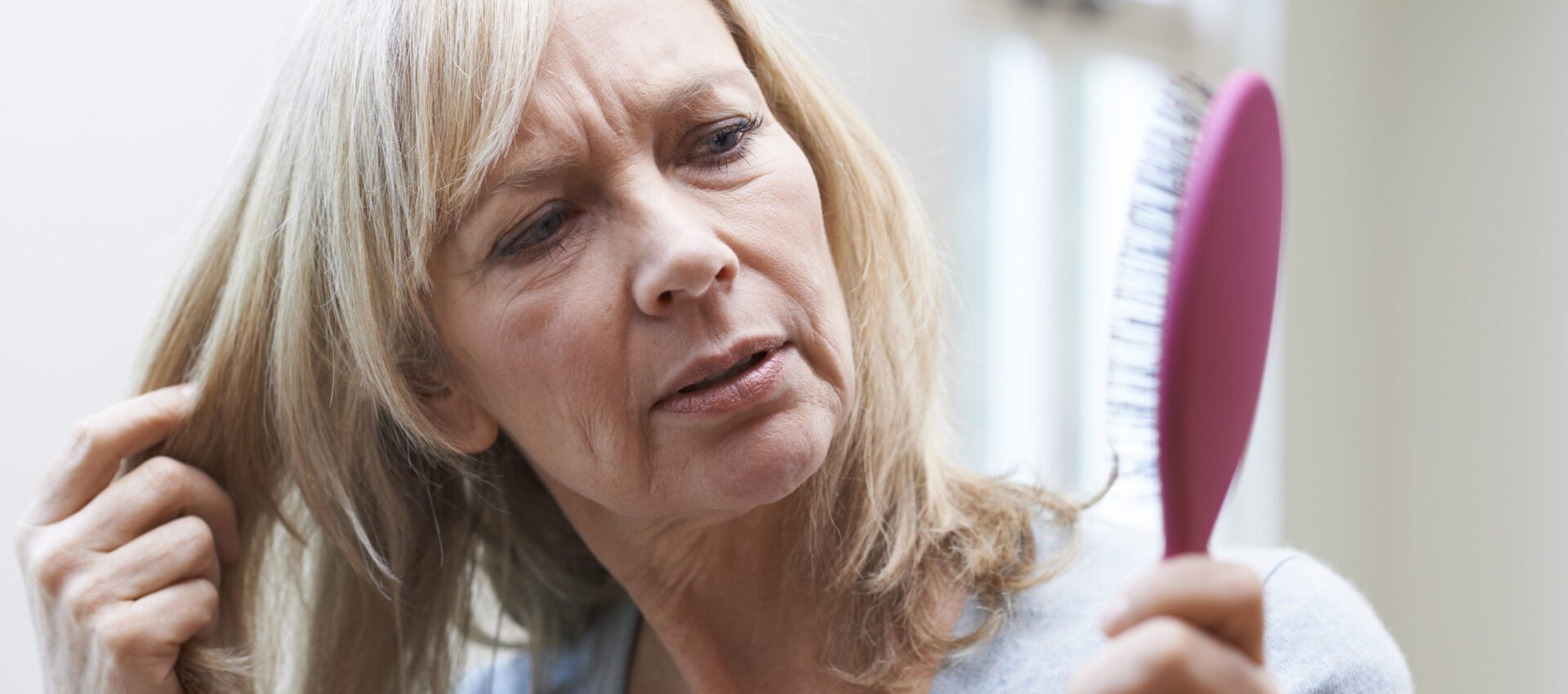A middle-aged woman looking concerned while examining her hair and holding a hairbrush with hair strands.