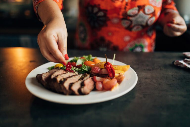 A person in a brightly colored shirt preparing a plate of food that includes sliced meat, vegetables, and garnishes, highlighting a balanced, protein-rich meal.