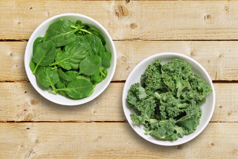 Two white bowls filled with fresh spinach leaves (left) and curly kale (right) placed on a wooden table.