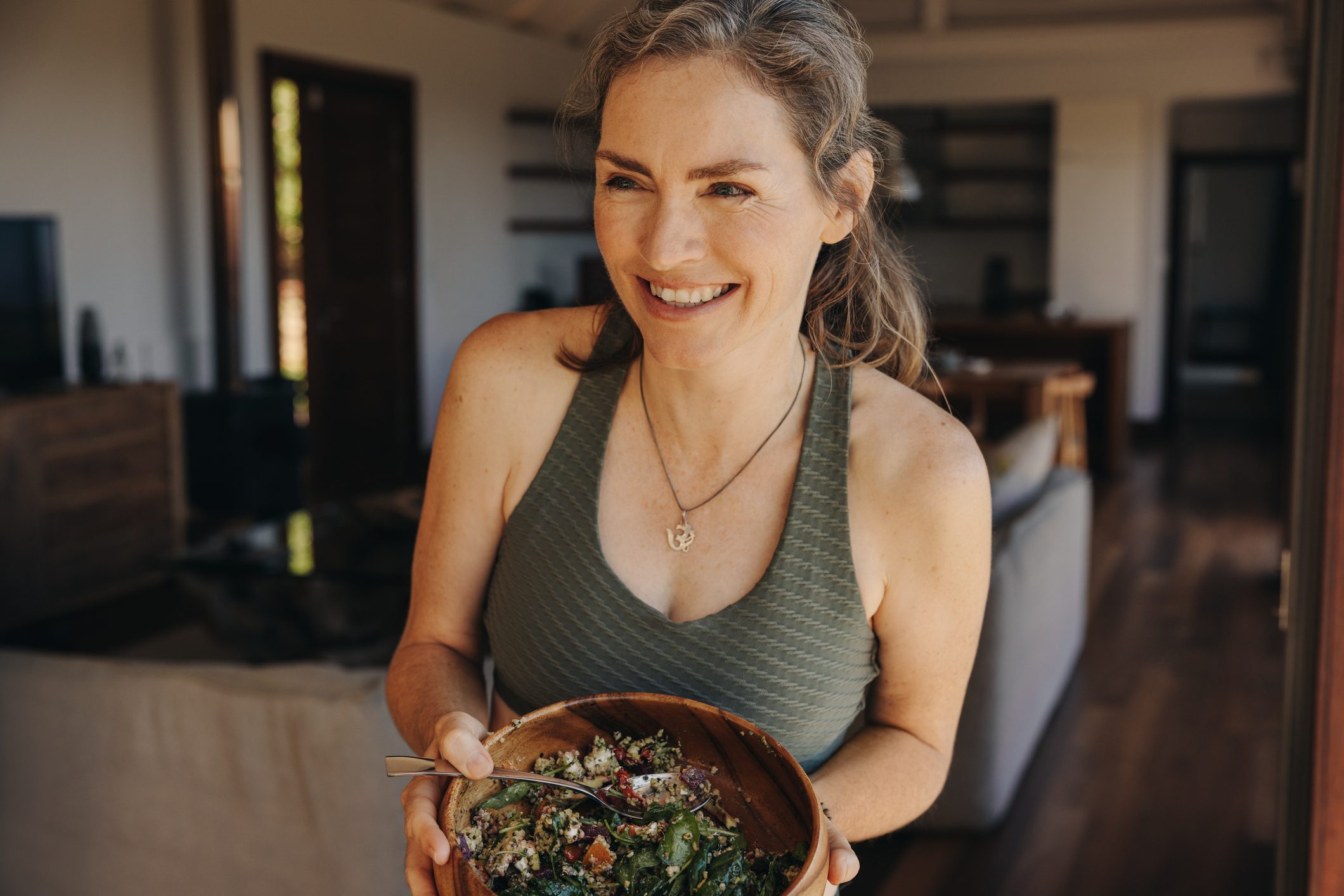 Smiling woman in a green tank top holding a wooden bowl filled with a fresh, healthy salad, standing in a cozy, well-lit home environment.
