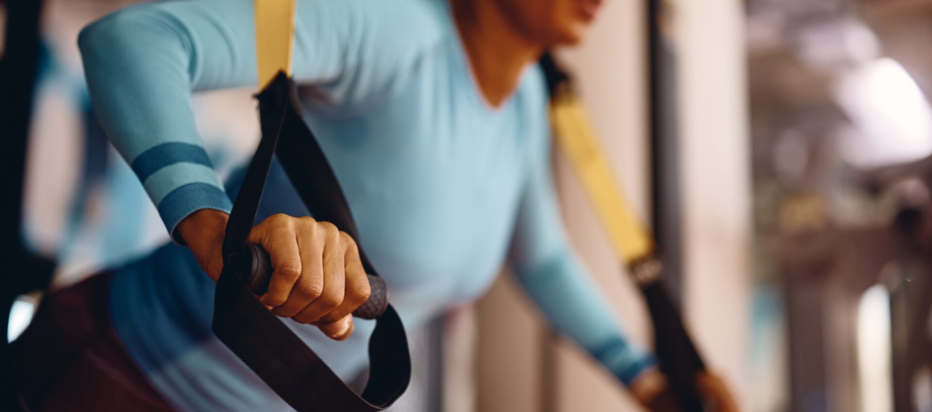 A woman in a blue long-sleeve shirt working out with TRX suspension bands in a gym, focusing intently while performing an exercise.