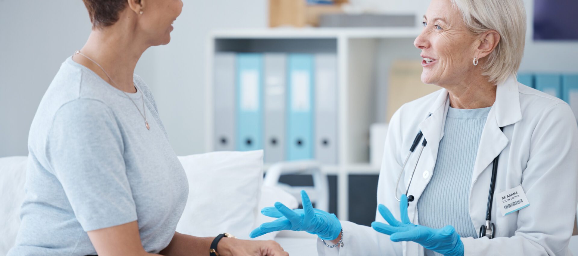 A female doctor in a white coat and gloves is having a discussion with a female patient, sitting on an examination table. The patient is attentively listening and appears to be engaged in the conversation. The setting is a modern medical office with organized files and medical equipment in the background.