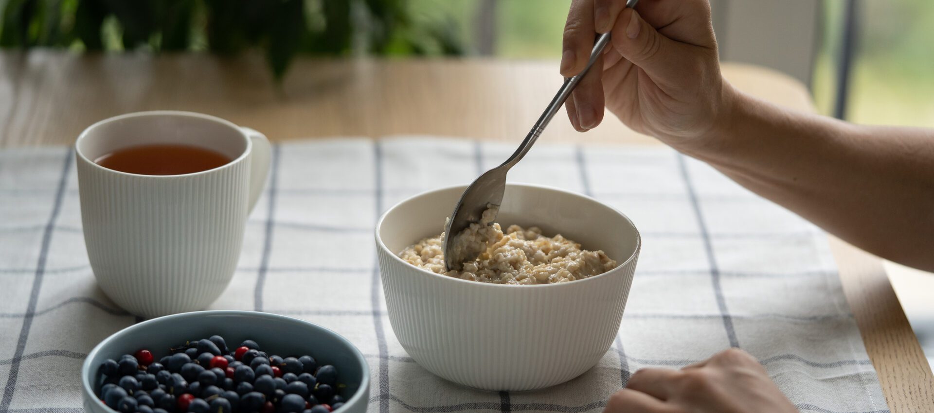 A healthy breakfast setup featuring a bowl of oatmeal being stirred with a spoon, accompanied by a bowl of mixed berries and a cup of tea, all set on a checkered tablecloth.
