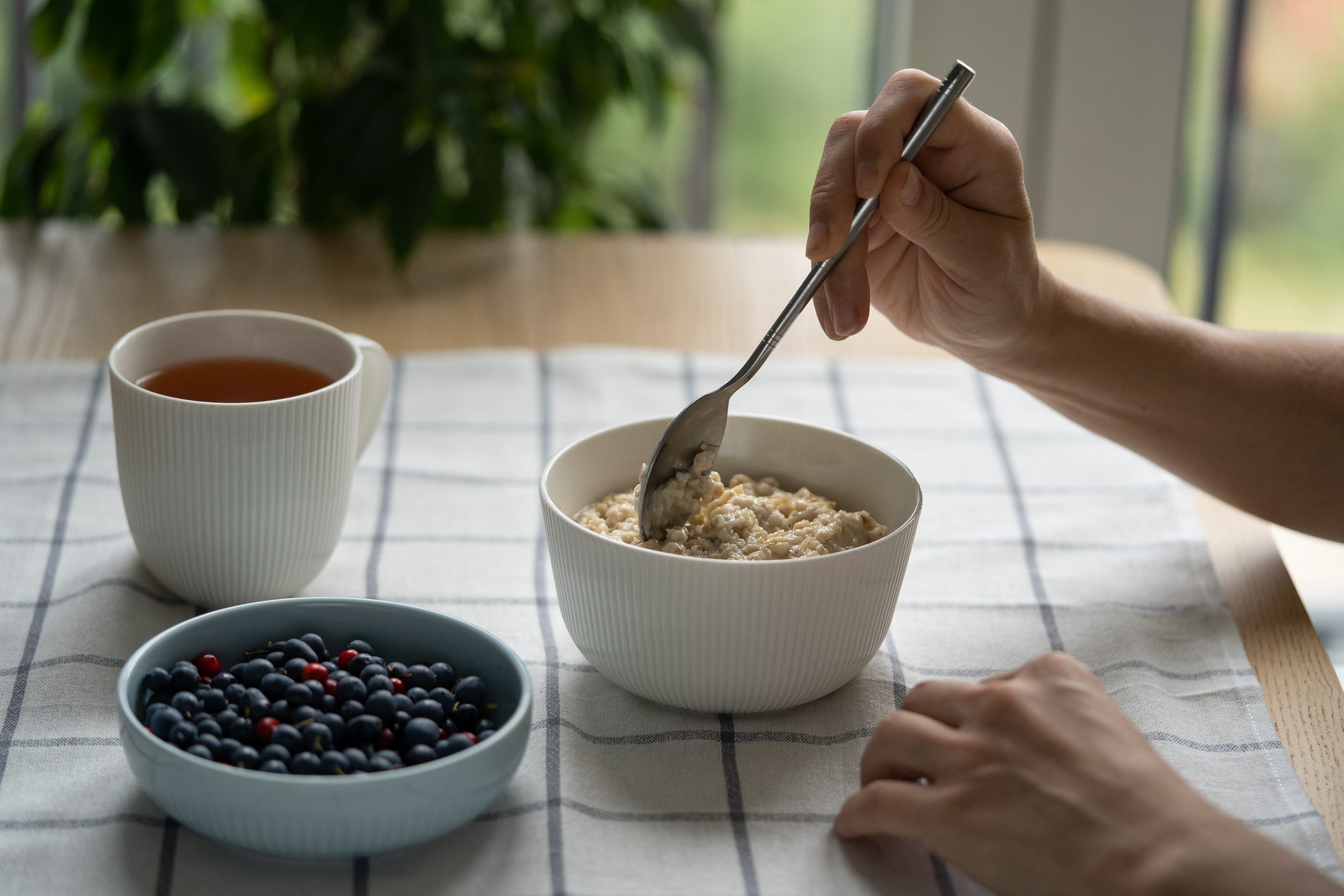 A healthy breakfast setup featuring a bowl of oatmeal being stirred with a spoon, accompanied by a bowl of mixed berries and a cup of tea, all set on a checkered tablecloth.