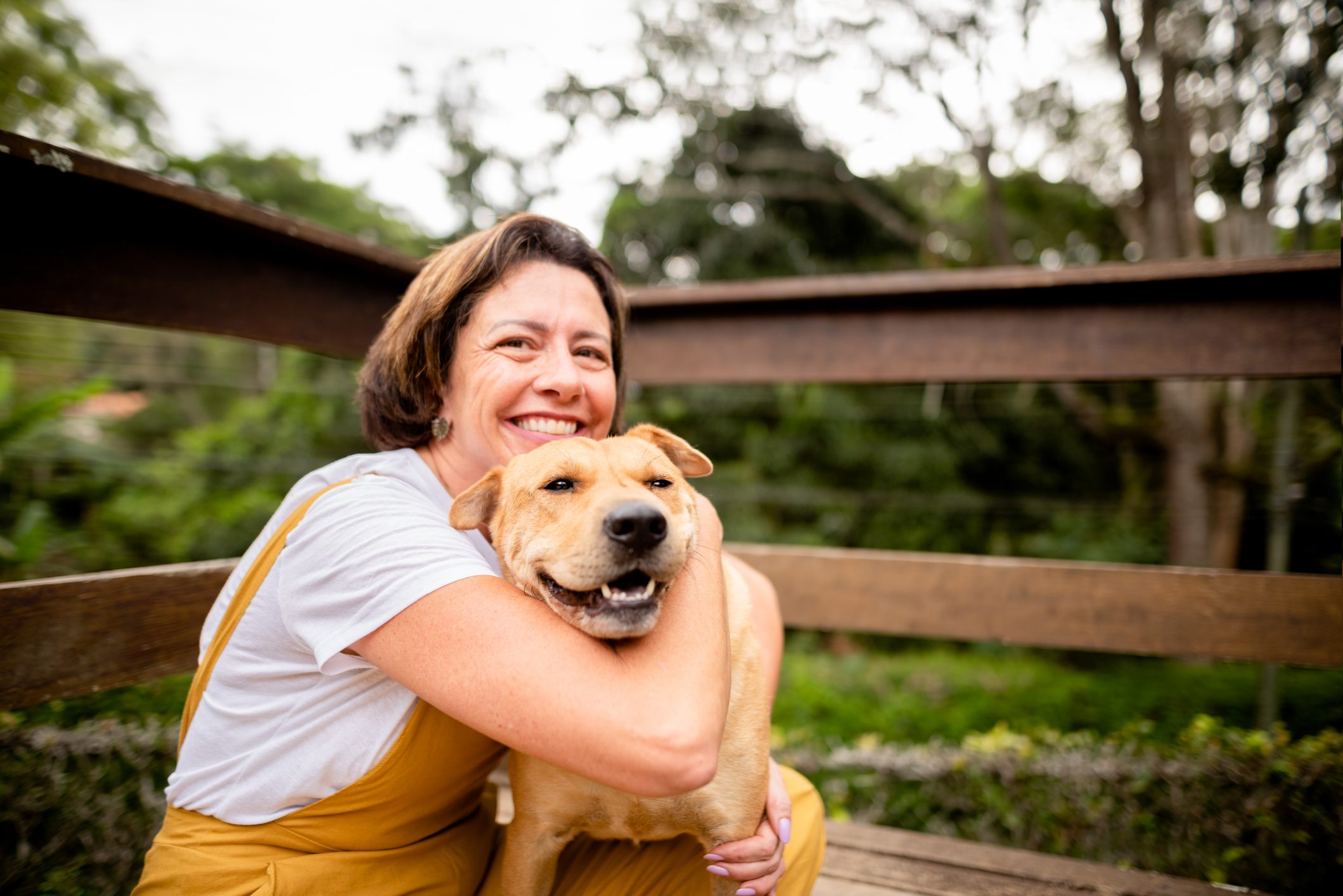 Smiling woman in a white t-shirt and yellow overalls hugging a happy dog on a wooden deck with a lush green background.