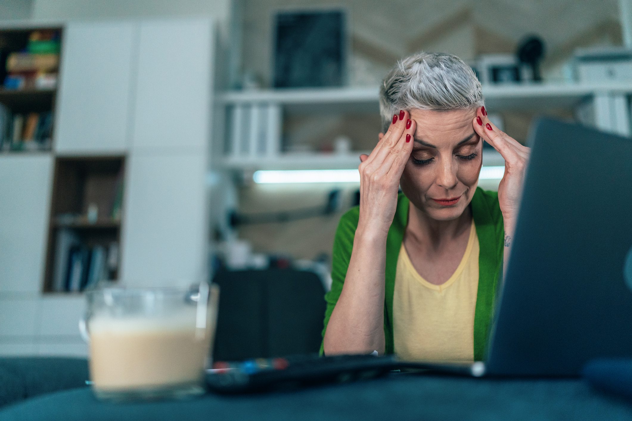 A woman with short gray hair, wearing a green cardigan and yellow shirt, sits in front of a laptop holding her temples with a stressed expression, illustrating the impact of menopause on daily life.