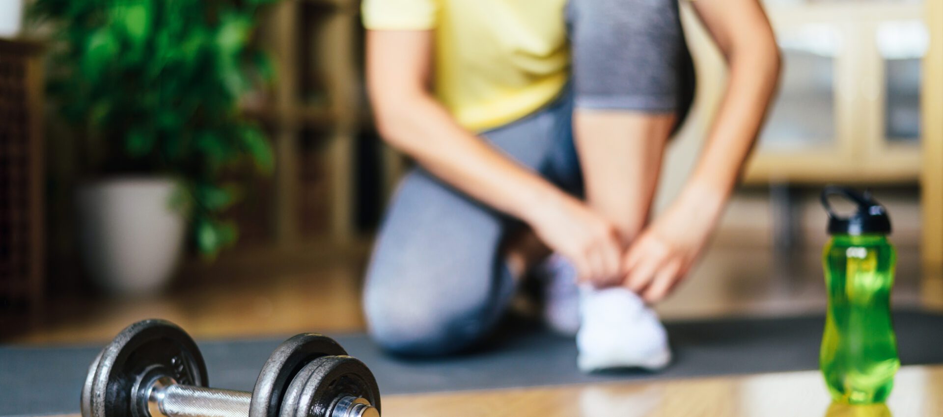 Person in workout attire tying their shoes on a yoga mat, with a dumbbell and water bottle in the foreground, preparing for a workout session.