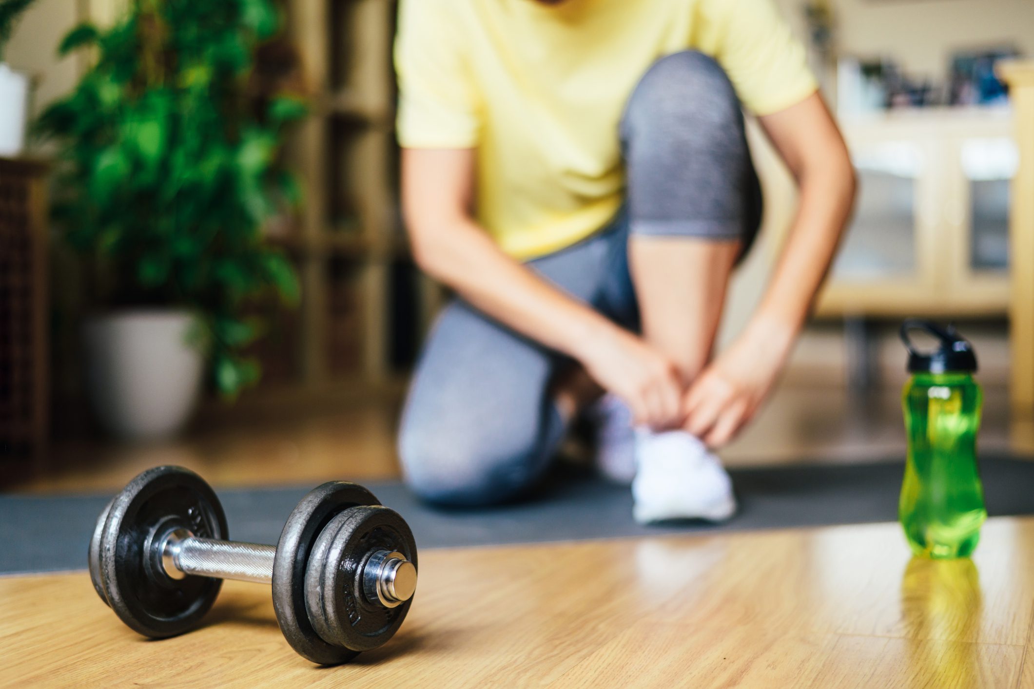 Person in workout attire tying their shoes on a yoga mat, with a dumbbell and water bottle in the foreground, preparing for a workout session.