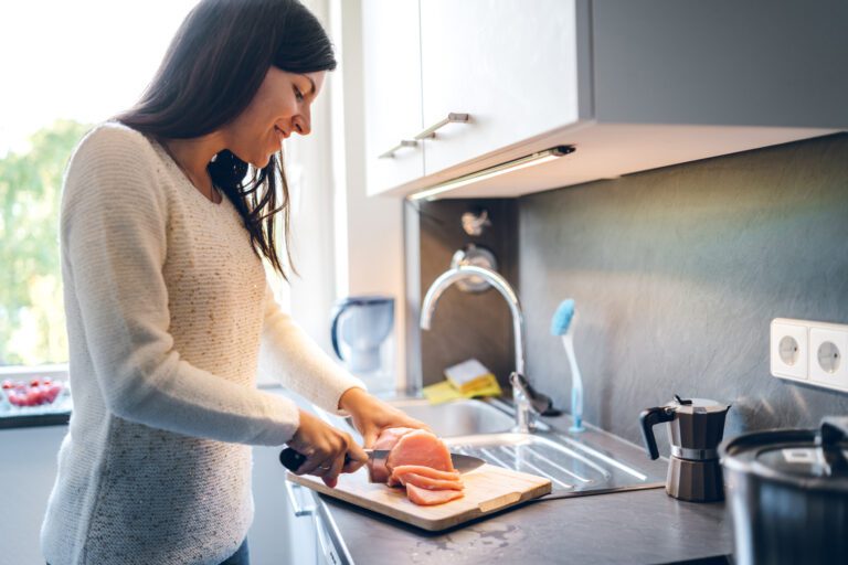 A woman in a white sweater preparing chicken in a kitchen, slicing the meat on a wooden cutting board near a sink with a smile on her face.