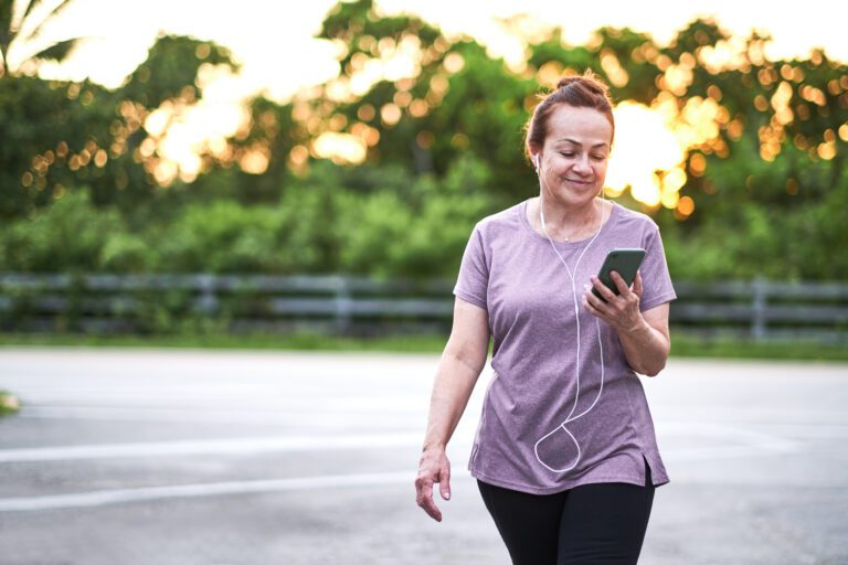 A middle-aged woman walking outdoors, looking at her phone with earphones in, wearing a purple shirt and black pants, with trees and sunset in the background.