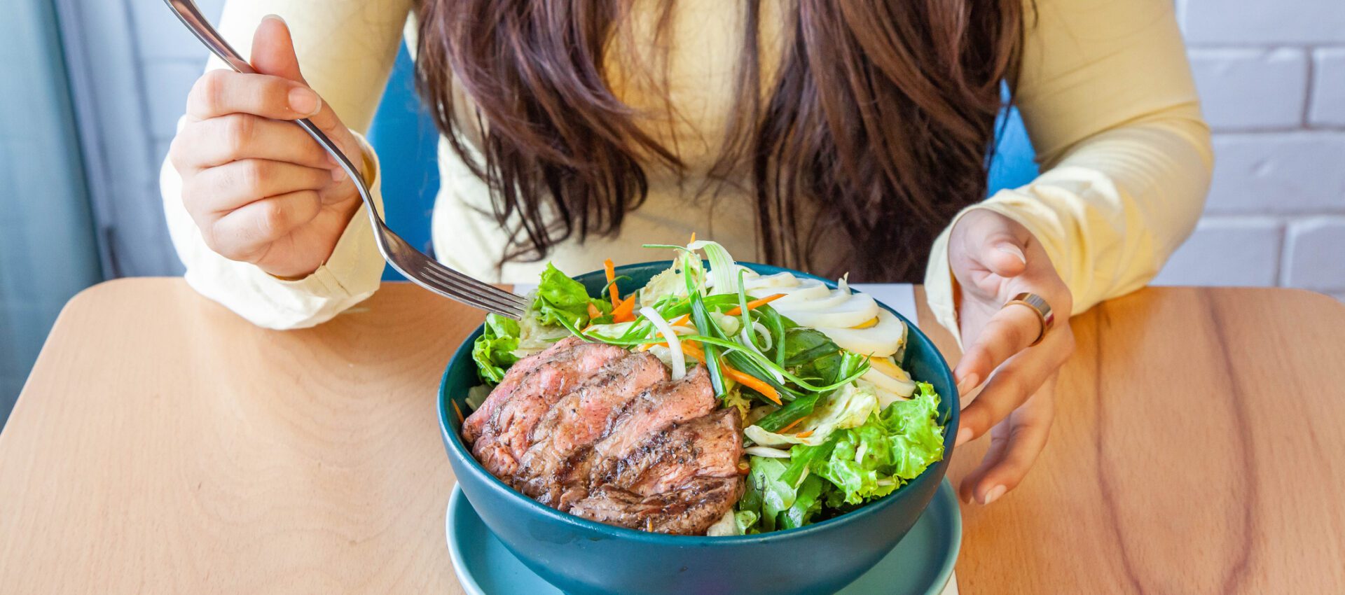 Woman enjoying a nutritious salad with grilled meat, leafy greens, and sliced vegetables, sitting at a wooden table in a casual dining setting.