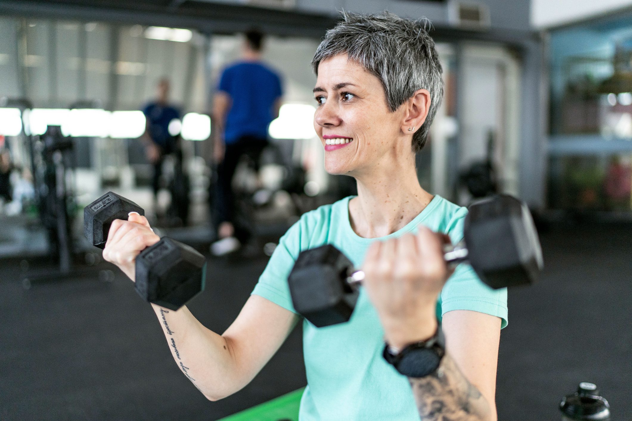 : A middle-aged woman with short gray hair smiling while lifting dumbbells during a strength training session at the gym. She is wearing a light blue t-shirt and has a tattoo on her forearm.