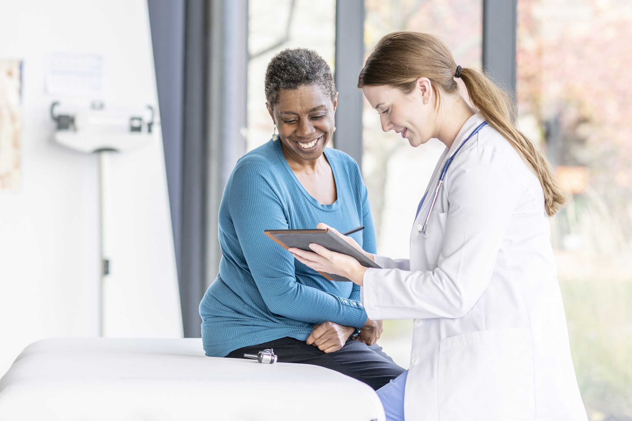 A woman in a teal sweater sits on an examination table, smiling and looking at a tablet held by a female doctor in a white coat, during a consultation.