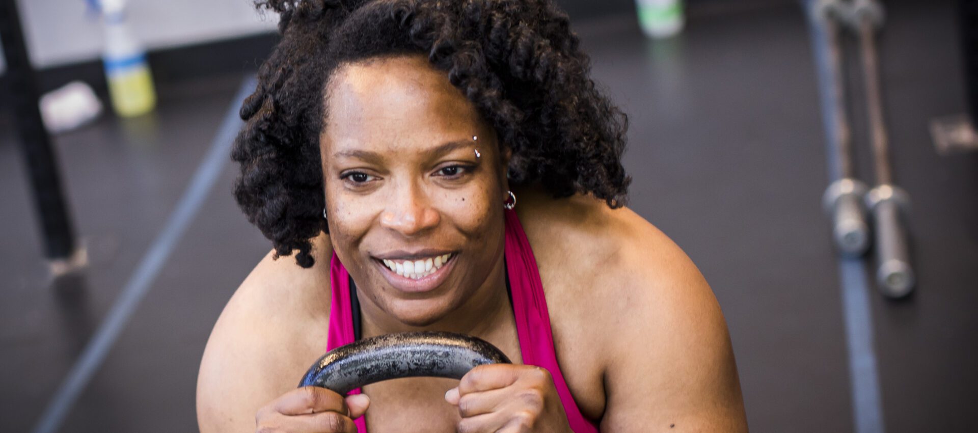 A woman with curly hair smiles and grips a kettlebell during a workout, demonstrating strength and determination.
