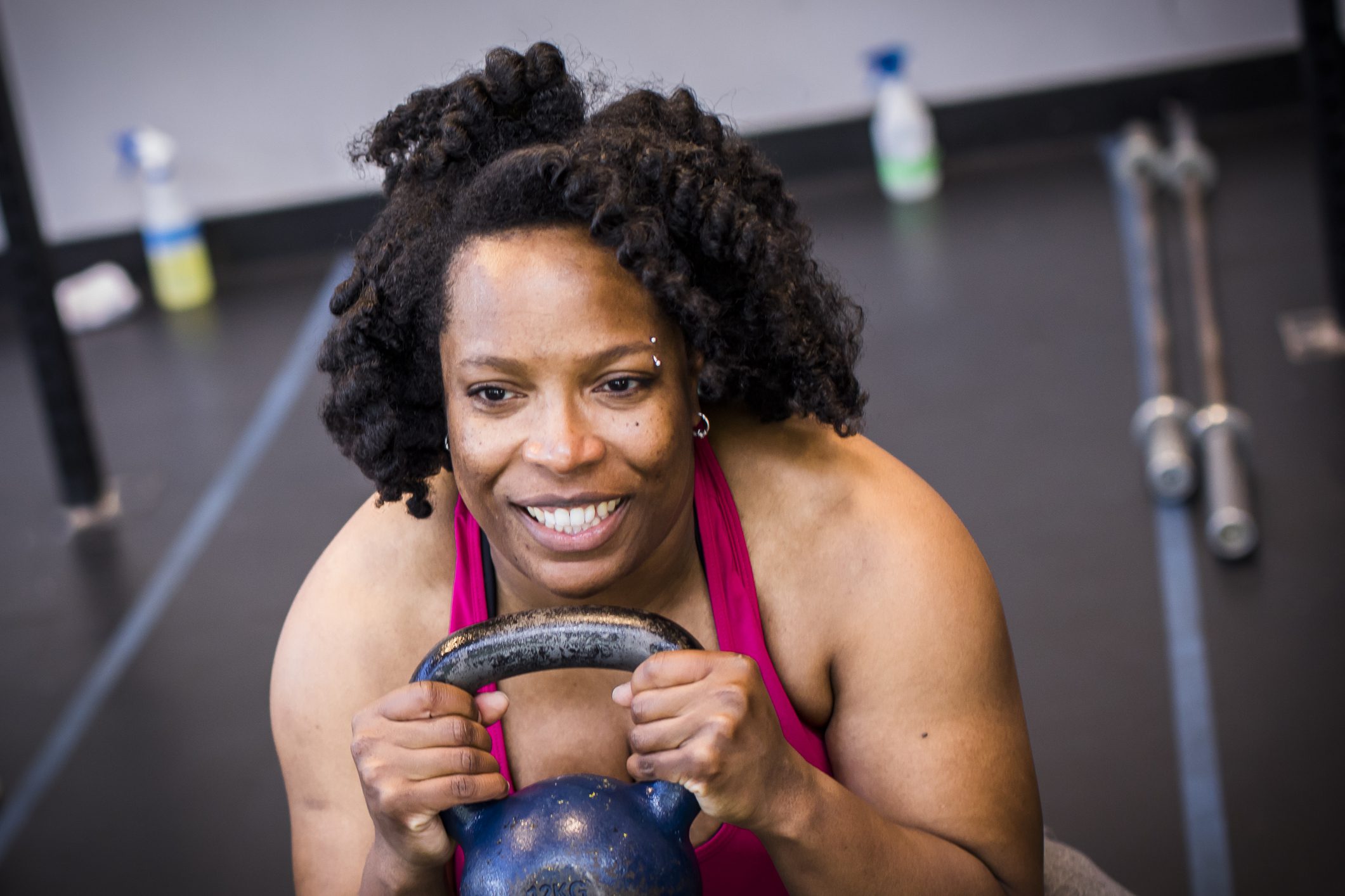 A woman with curly hair smiles and grips a kettlebell during a workout, demonstrating strength and determination.