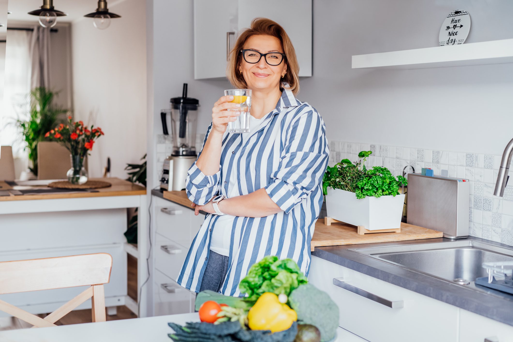 A woman with short blonde hair and glasses, wearing a striped shirt, stands in a modern kitchen holding a glass of lemon water. Fresh vegetables and herbs are visible on the counter in front of her, and the kitchen is bright and well-lit.