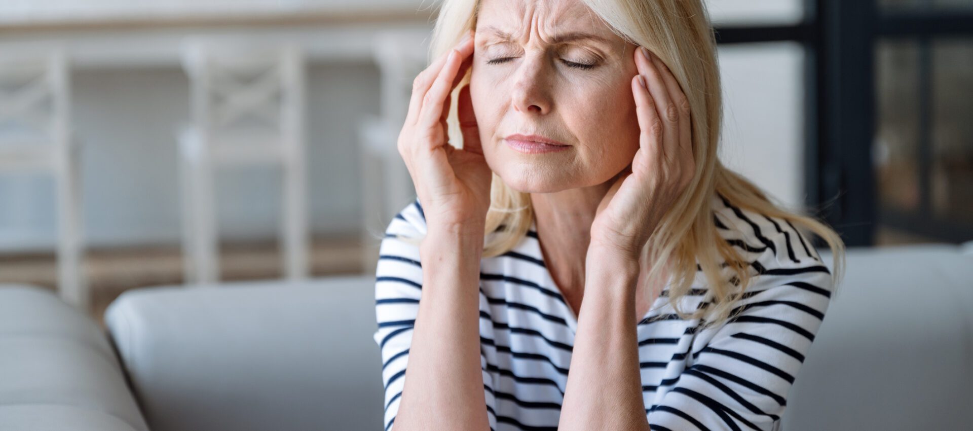A middle-aged woman with blonde hair, sitting indoors and holding her head with both hands, appearing to suffer from a headache or stress.