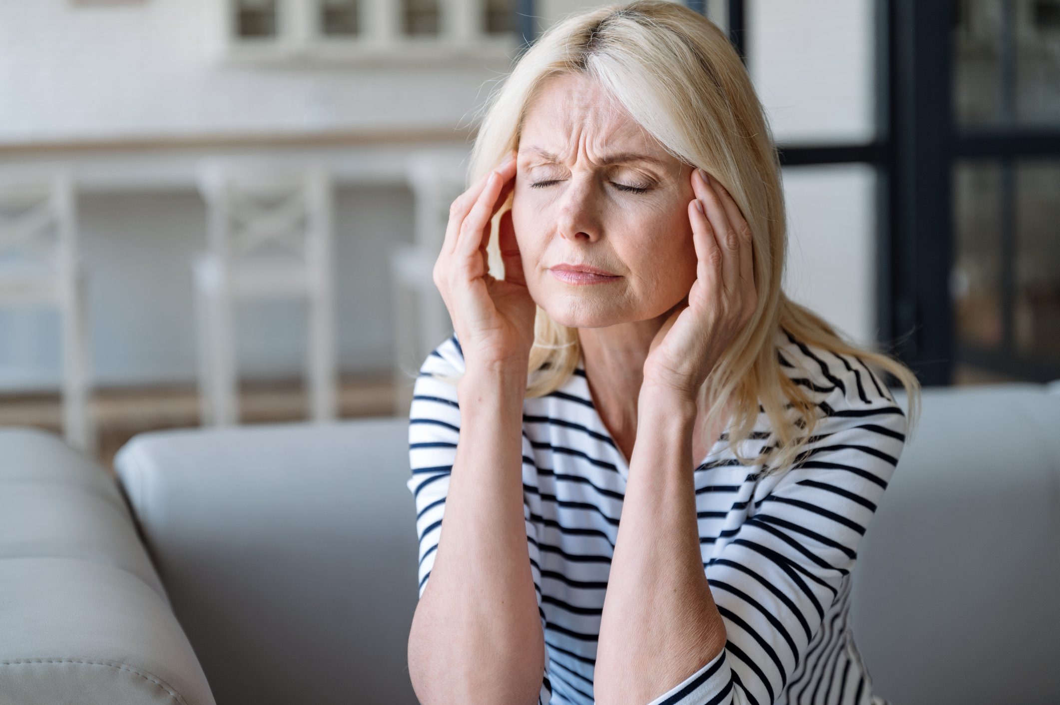 A middle-aged woman with blonde hair, sitting indoors and holding her head with both hands, appearing to suffer from a headache or stress.