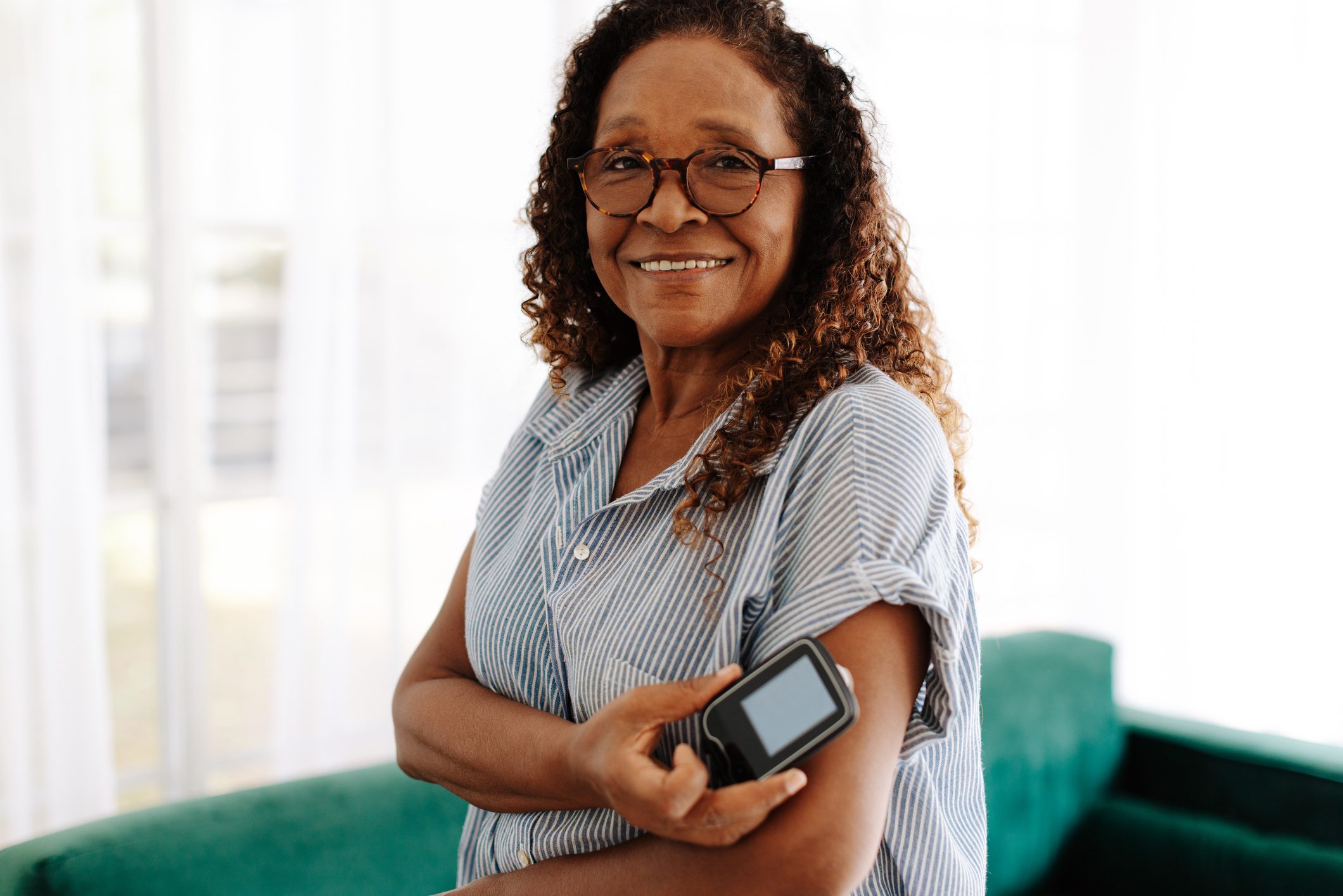 A smiling woman with curly hair and glasses, wearing a striped shirt, uses a glucose monitor on her arm. She is standing in a bright room with a green couch in the background.