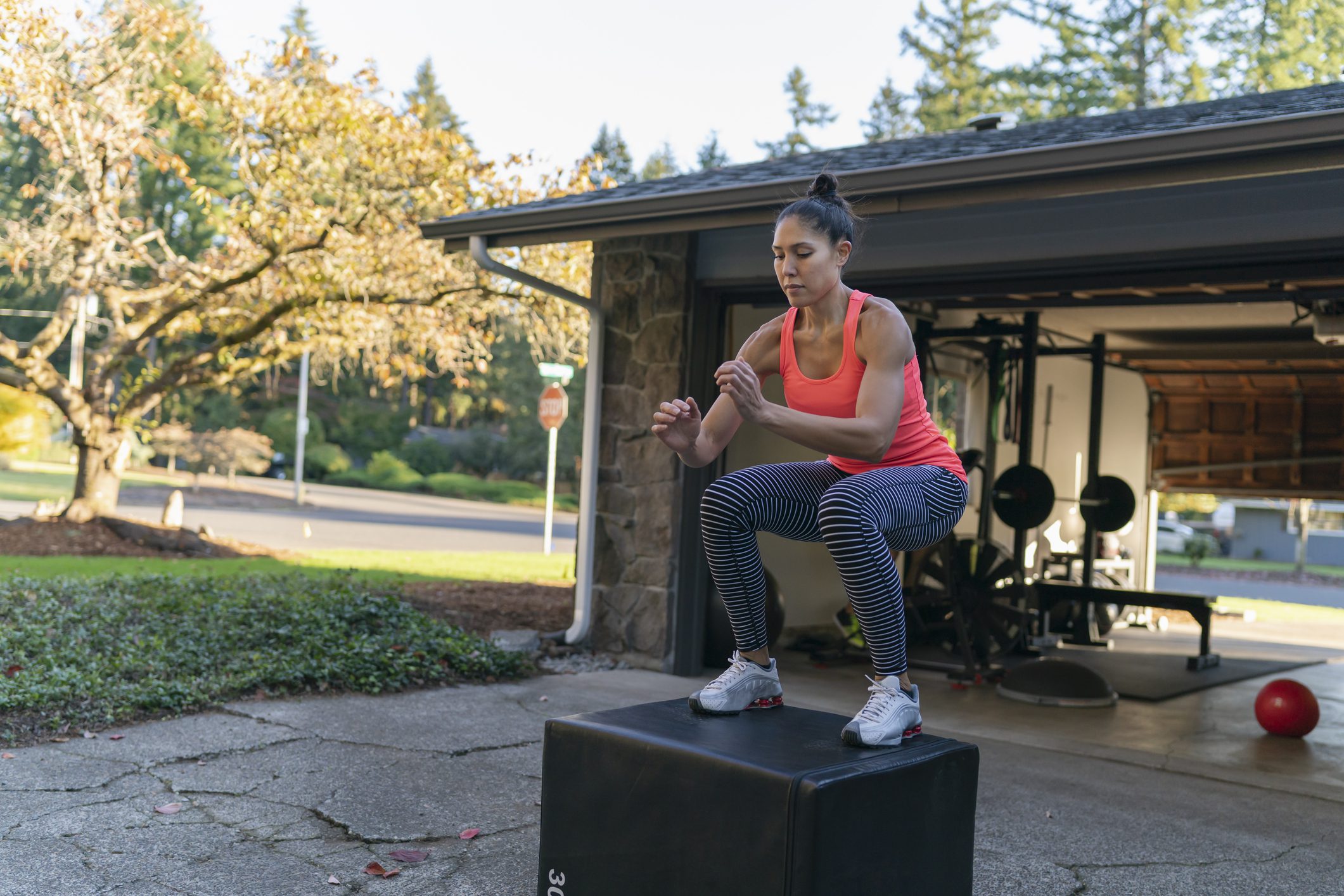 Woman performing box jumps outdoors in front of a garage gym, wearing a bright pink tank top and striped leggings. She is mid-jump with knees bent, arms engaged, and focused expression, demonstrating an athletic workout routine.