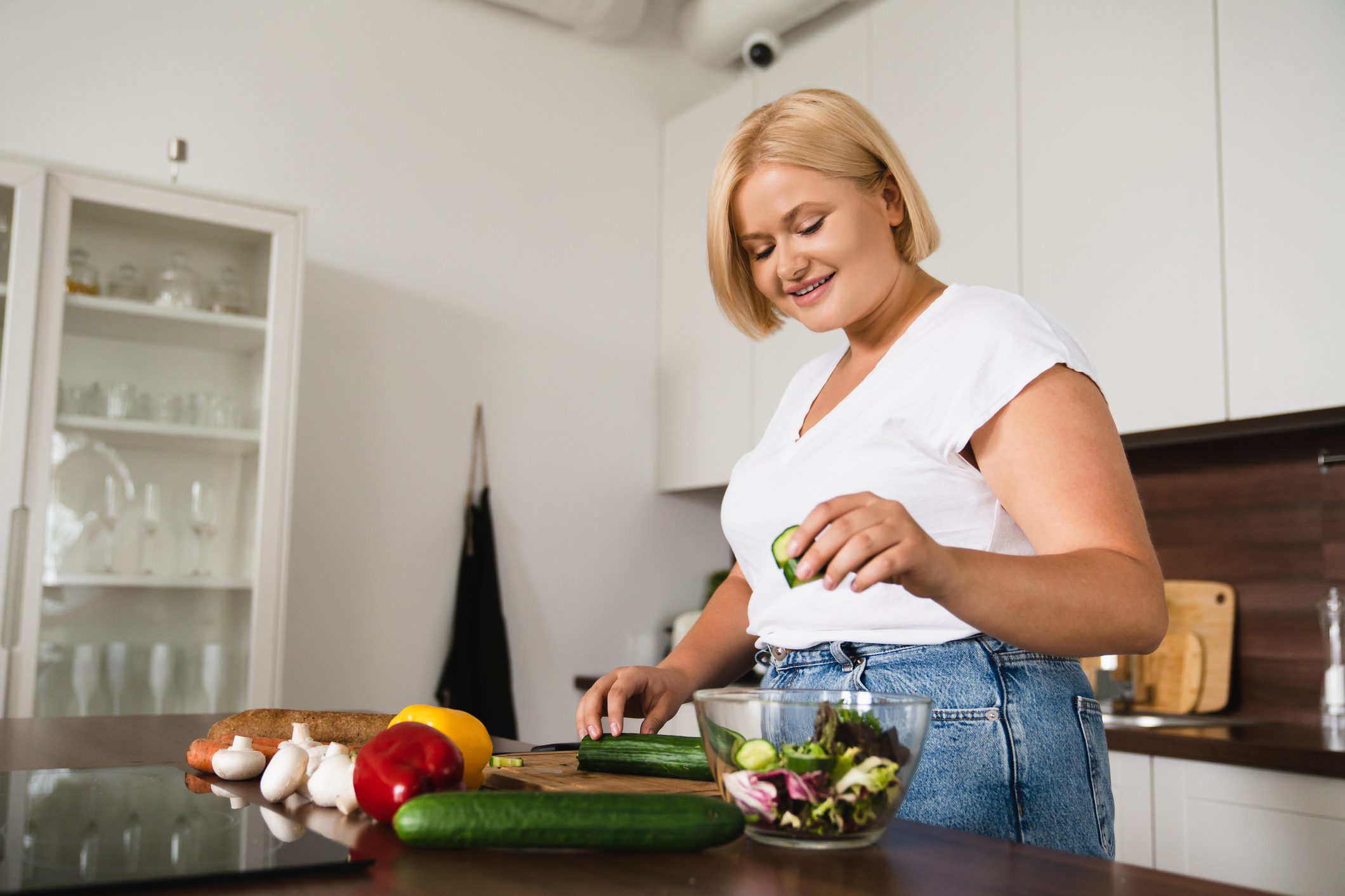 A woman with blonde hair and a white shirt smiles as she prepares a fresh salad in her kitchen, surrounded by a variety of colorful vegetables on the countertop.