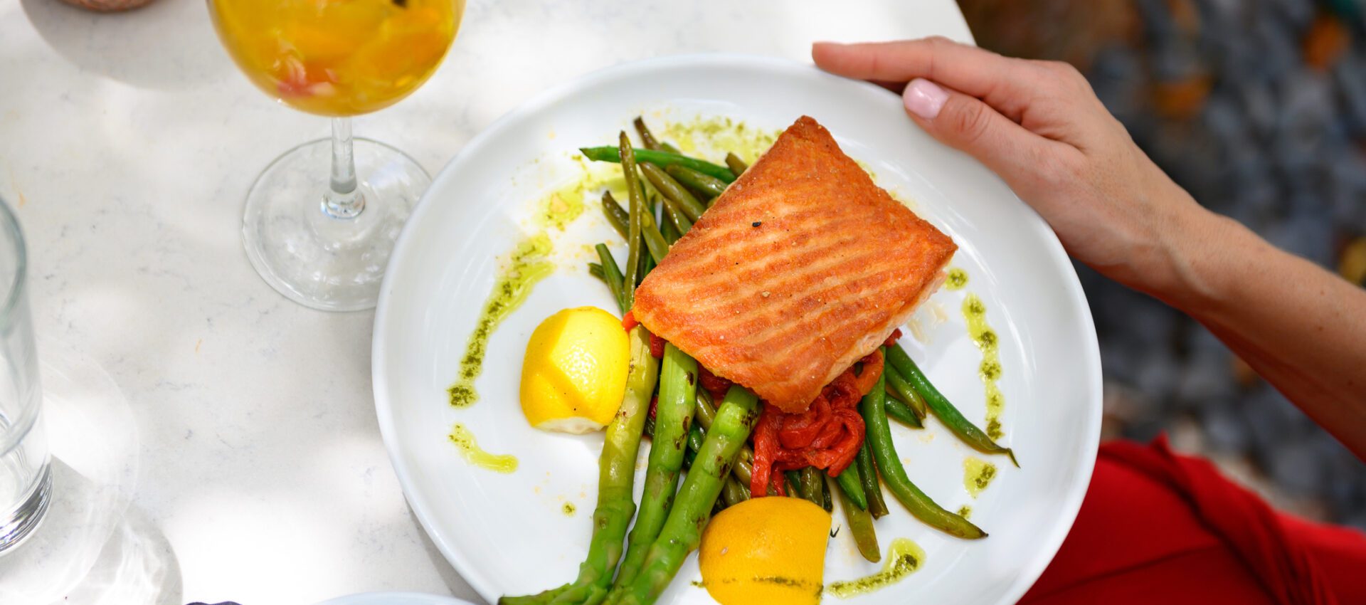 A plate of grilled salmon served on a bed of green beans and asparagus, garnished with lemon wedges and a drizzle of pesto sauce. The dish is accompanied by a refreshing iced beverage on a white table, creating a vibrant and healthy meal setting.
