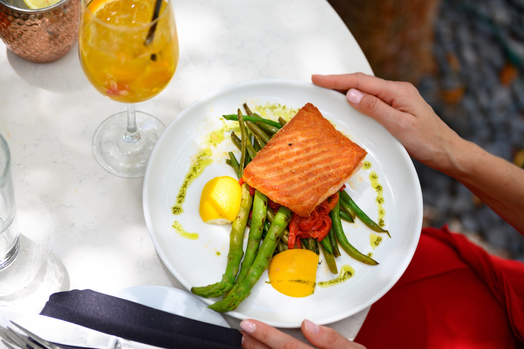 A plate of grilled salmon served on a bed of green beans and asparagus, garnished with lemon wedges and a drizzle of pesto sauce. The dish is accompanied by a refreshing iced beverage on a white table, creating a vibrant and healthy meal setting.