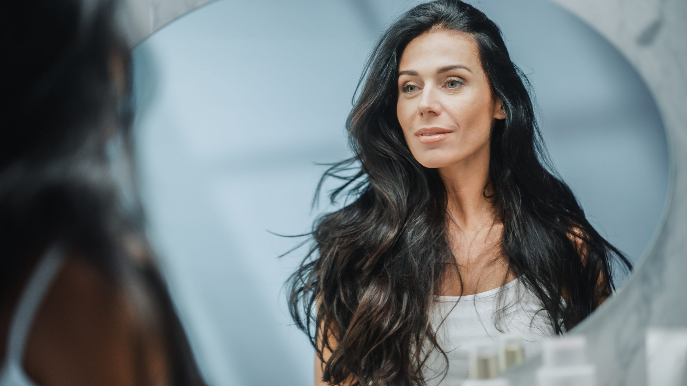 A woman with long, dark hair gazes thoughtfully into a mirror, her reflection showing a calm and introspective expression. The background is softly blurred, emphasizing her clear, healthy complexion.