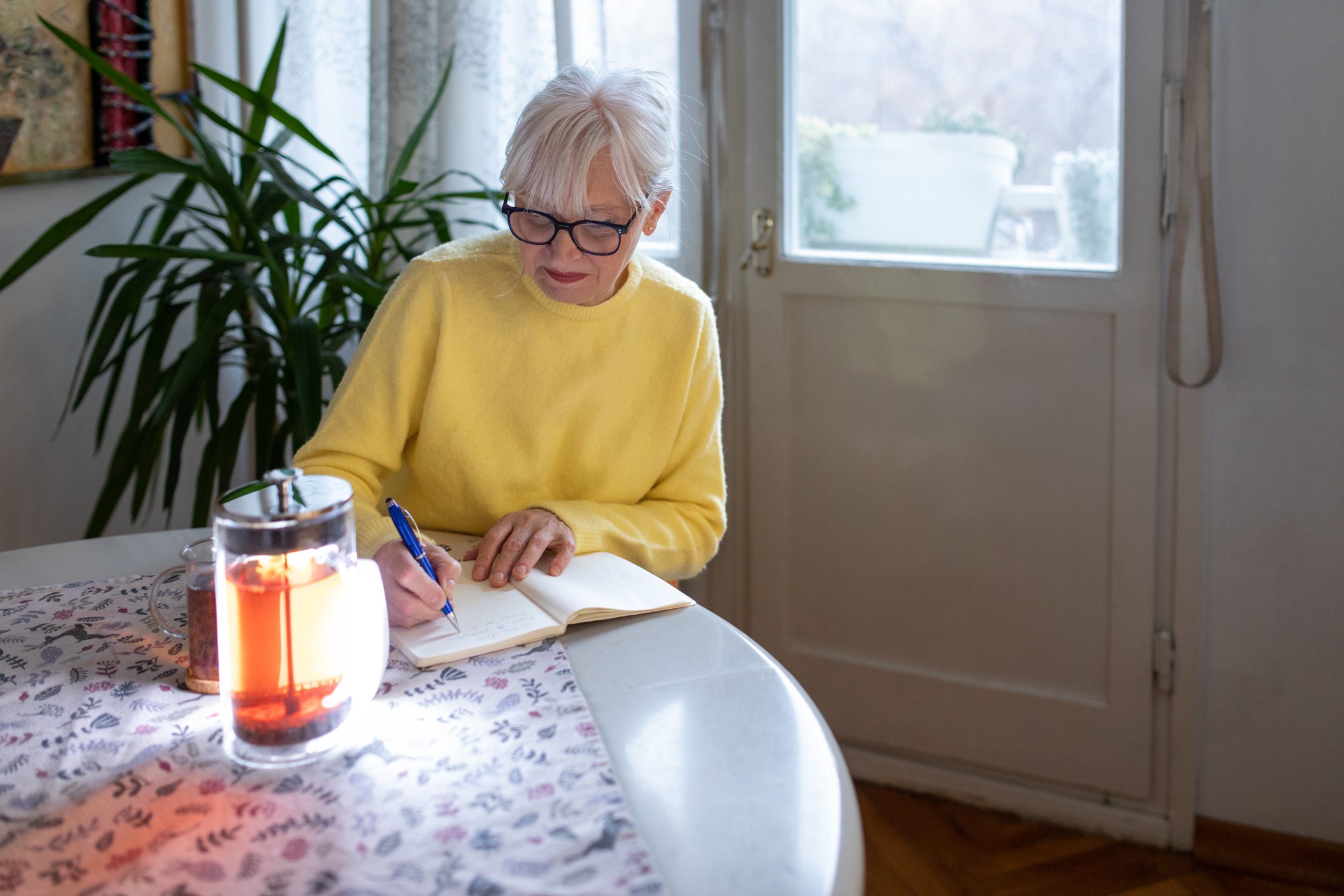 An older woman wearing glasses and a bright yellow sweater sits at a table, writing in a journal. A glass teapot filled with tea is on the table beside her, and sunlight streams through a nearby window, creating a peaceful and reflective atmosphere.