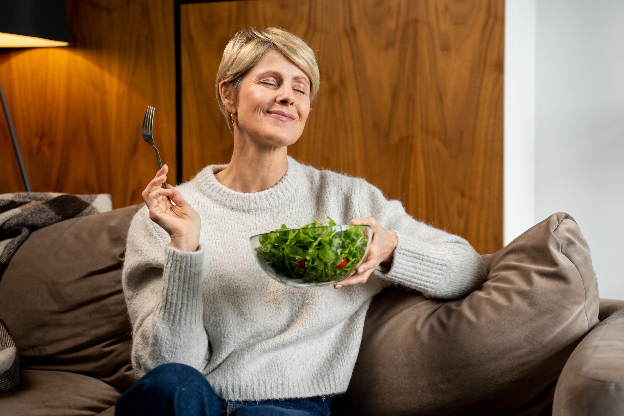 A woman with short blonde hair smiles contentedly while holding a bowl of fresh salad and a fork, savoring the healthy meal on her cozy couch.