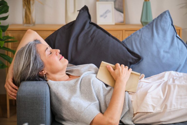 Woman with gray hair relaxing on a couch, holding a book, eyes closed, enjoying a peaceful moment.