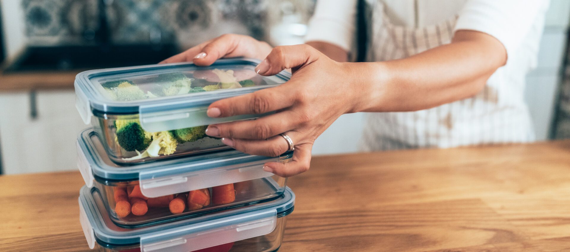 A person in a kitchen stacking glass meal prep containers filled with chopped vegetables, including broccoli, carrots, and tomatoes. The image emphasizes the organized and efficient preparation of healthy meals in advance.
