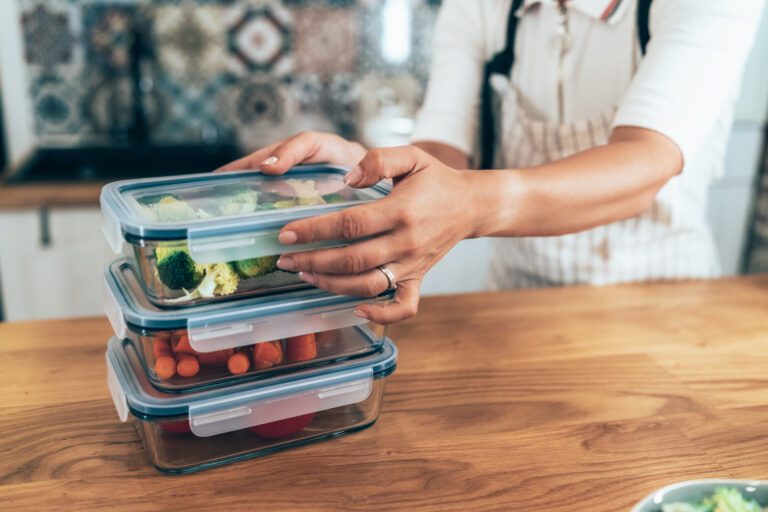 A person in a kitchen stacking glass meal prep containers filled with chopped vegetables, including broccoli, carrots, and tomatoes. The image emphasizes the organized and efficient preparation of healthy meals in advance.