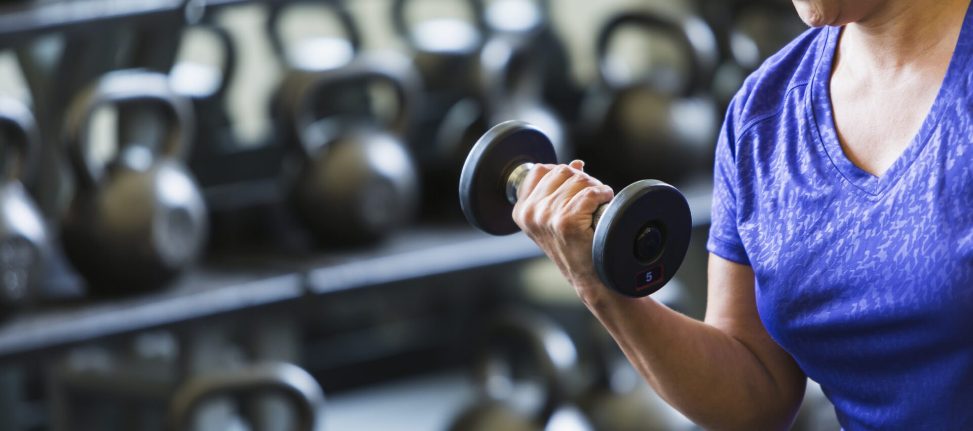 A woman in a purple shirt lifting a 5-pound dumbbell in a gym, focusing on strength training. In the background, there are several kettlebells lined up on racks, emphasizing a fitness environment dedicated to resistance training.