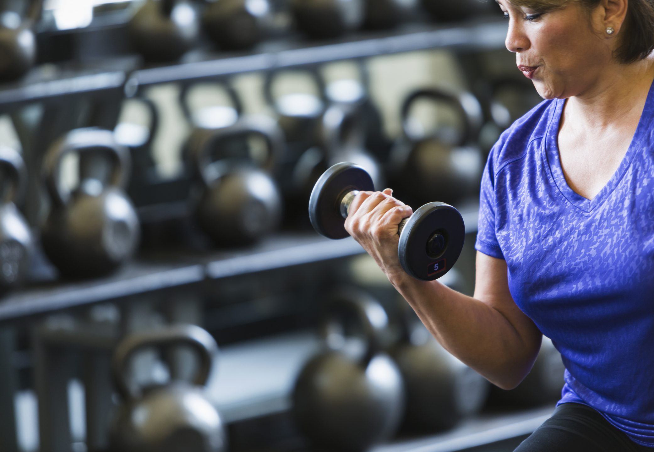 A woman in a purple shirt lifting a 5-pound dumbbell in a gym, focusing on strength training. In the background, there are several kettlebells lined up on racks, emphasizing a fitness environment dedicated to resistance training.